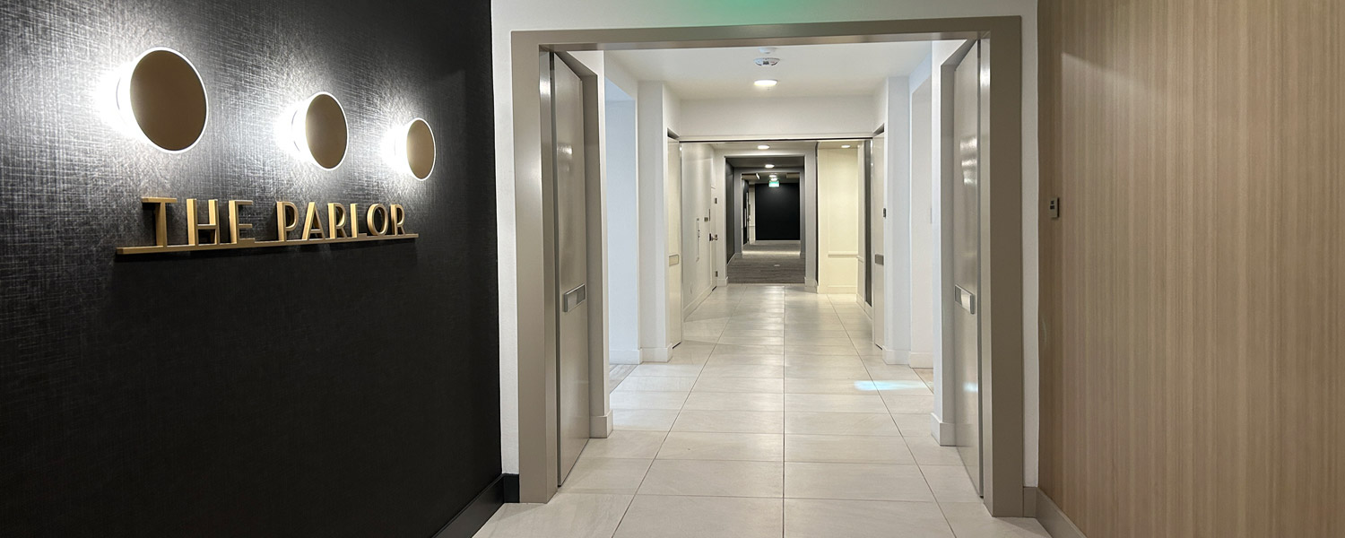 Syntegra-Area-Separation-Doors Hotel corridor Area Separation Door system with elevators on both sides, a dark accent wall with three circular lights and gold 'THE PARLOR' sign.