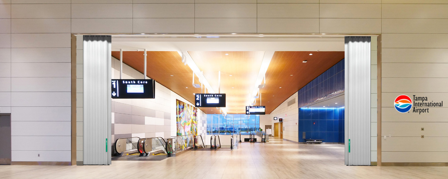 Wondoor-FireGuard-Tampa-Airport Airport concourse at Tampa International Airport showing the Won-Door FireGuard™ Accordion System, escalators, signboards, and a bright, modern waiting area.